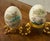This pair of vintage decorative eggs showcases delicate hand-painted artistry, each resting on an ornate gold tone metal...