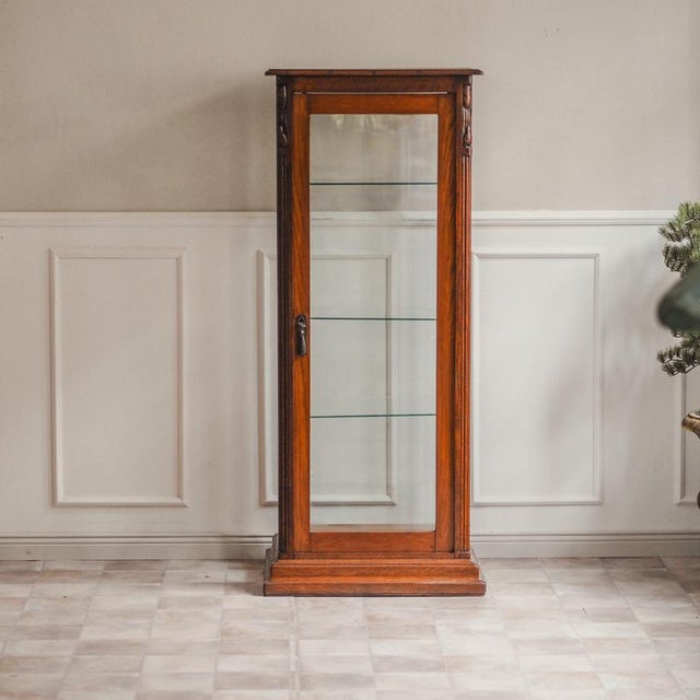 A narrow, three-sided glazed oak display cabinet, created around 1900 during the transition from historicist architectural...