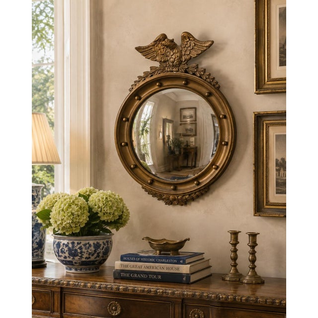 A Federal-style giltwood convex mirror crowned with a carved eagle, its wings outstretched above a laurel motif—an...