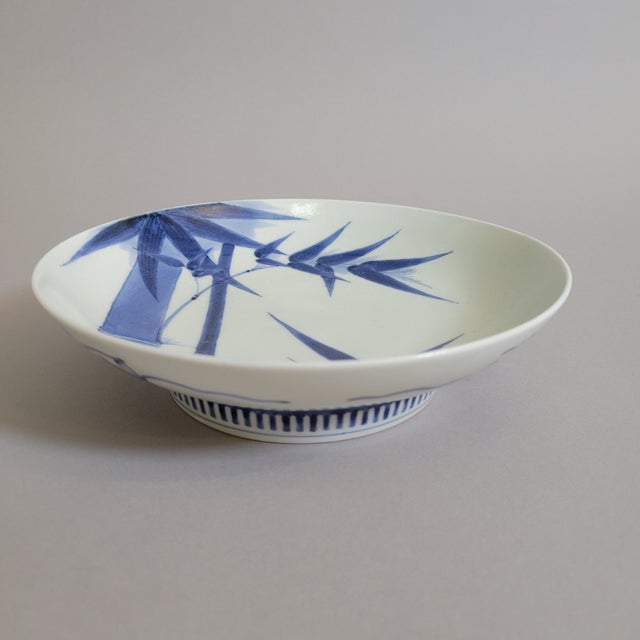 A refined Japanese porcelain shallow bowl featuring a hand-painted bamboo motif in underglaze cobalt. The composition is...