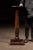 Solid wood pedestal stand featuring a circular top over a tapered, turned column rising from a square plinth base. The...