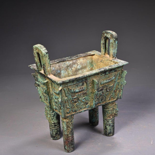 This is a cast bronze rectangular vessel, featuring a sturdy form with two upright loop handles and four cylindrical legs....