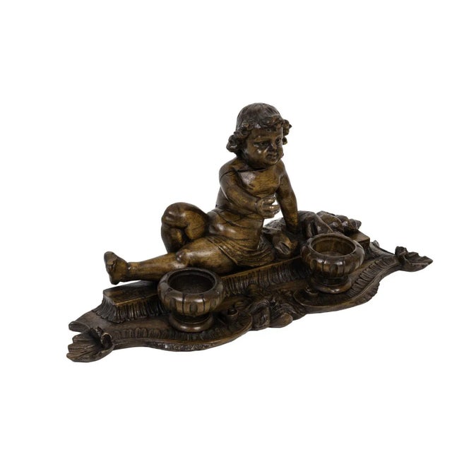 Inkwell in wood figuring a slightly lied down child only dressed with a simple cloth around his hips. He stands on a small...