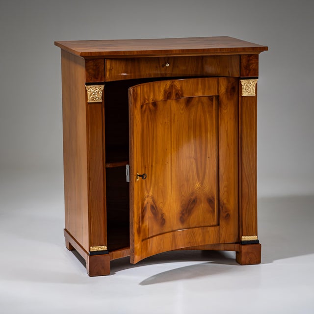 Biedermeier demi-cabinet in cherrywood with a concave front and flanking pilasters featuring gold-patinated capitals and...
