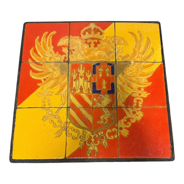 A striking vintage accent table featuring a vivid enamel-tiled top emblazoned with an ornate European heraldic crest. The...