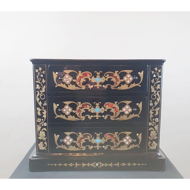 This black polished wood commode is decorated with brass stringing on its top. The front is inlaid with brass leaf...