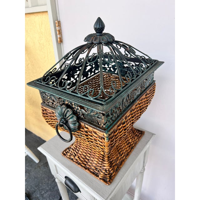 This item is a decorative lidded wicker storage basket featuring a wrought iron cage lid with a grape leaf motif. The...