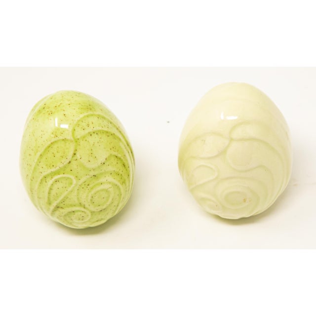 Offering a pair of ceramic glazed eggs. One is green with speckles and a swirled pattern. The other is solid cream with...