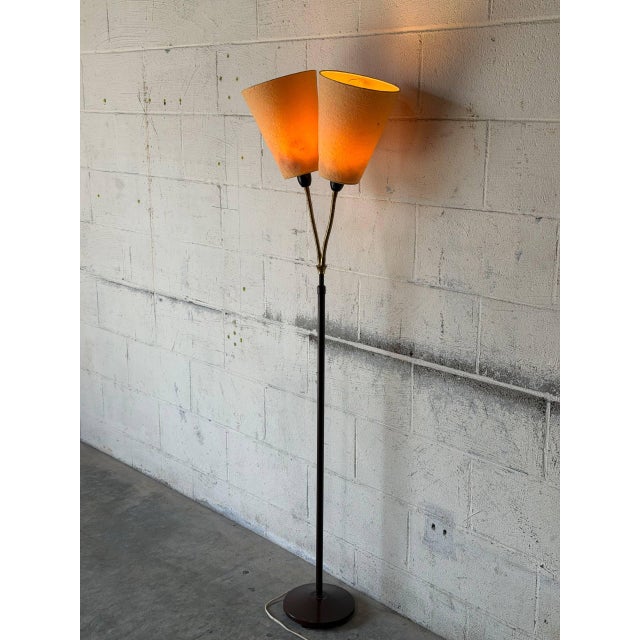 This striking floor lamp features a slender dark metal stem branching into dual adjustable arms with textured cone shades....