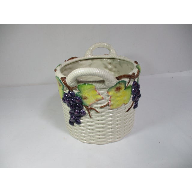 A large white glazed ceramic woven basket with handles. It is decorated with raised purple grapes and leaves in shades of...