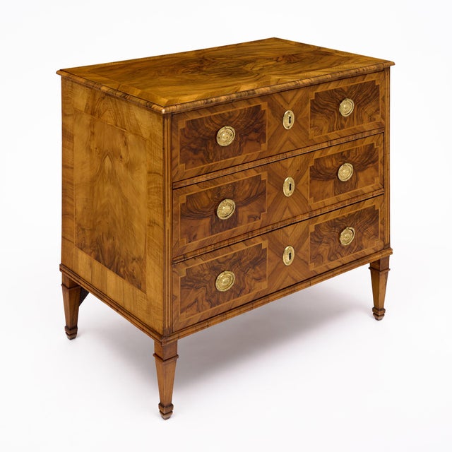 An elegant three-drawer commode showcasing beautifully figured walnut veneers arranged in a sophisticated symmetrical...