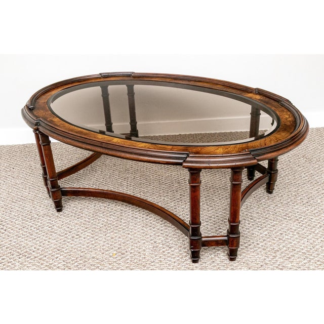 A vintage Regency Revival oval coffee table, circa 1970s, with a richly figured wood frame and an inset beveled glass top....