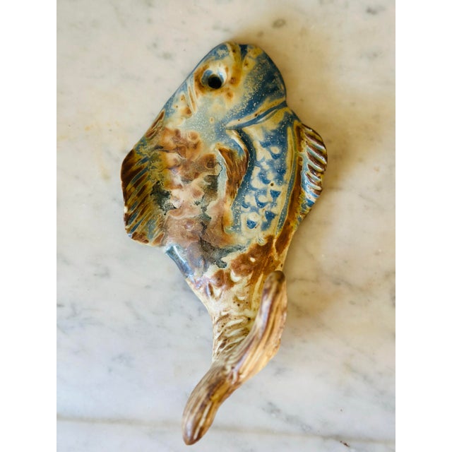 A handcrafted ceramic wall hook featuring an abstract fish form with a sculptural curved hook and richly mottled coastal...