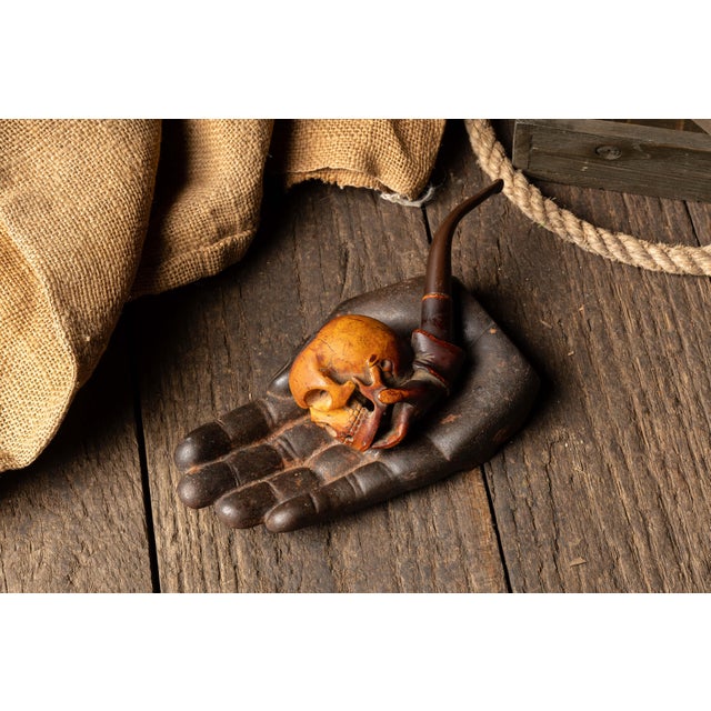 Carved figural pipe depicting a skull cradled in an outstretched hand, paired with a matching sculpted hand tray. Executed...