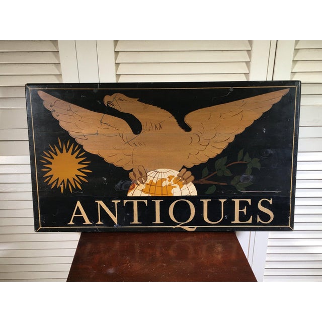 A hand-painted wooden trade sign with a golden folk art style eagle with outstretched wings gripping a globe, radiant sun...