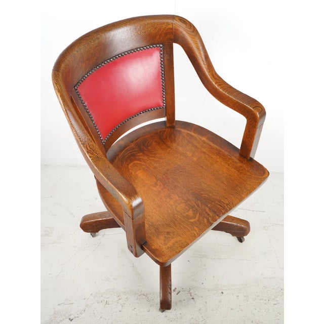Early 20th century swivel office chair made of solid oak with a studded red vinyl backrest. It features an adjustable...