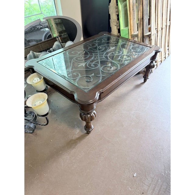A showpiece coffee table featuring a heavy dark wood frame with ornately carved legs and a glass top that reveals...