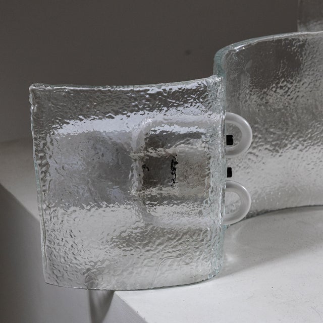 An elegant pair of wall sconces featuring thick, curved glass diffusers with a refined hammered finish, creating soft,...