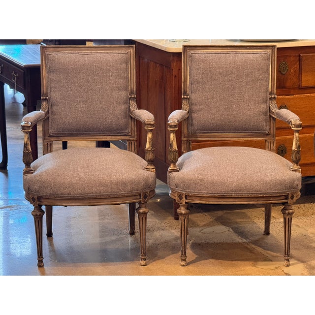 A pair of Louis XVI style armchairs in painted gray finish with carved fluted legs and scrolled arms. Each chair features...