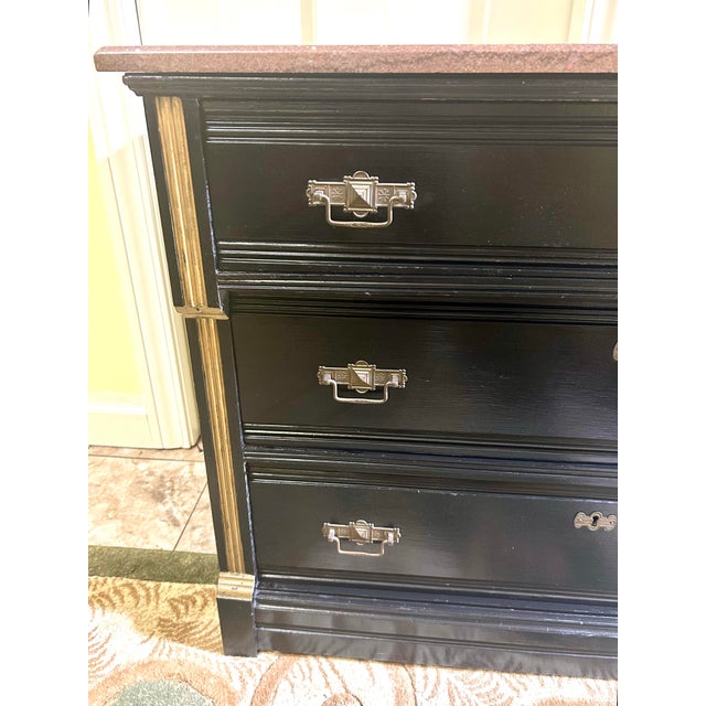 This vintage, ebonized-wood commode, features a marble top, three drawers with gilded bronze mounts, and gilded pilasters....