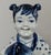 A charming vintage blue-and-white porcelain figurine depicting a smiling child in traditional dress, holding a large...