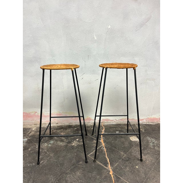 A charming pair of Mid-Century Modern bar stools featuring handwoven natural rattan tops supported by sleek black iron...