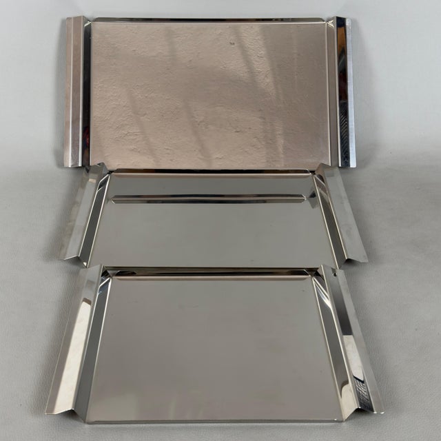 A simple and refined serving set consisting of three stackable rectangular stainless steel trays, authentically marked...