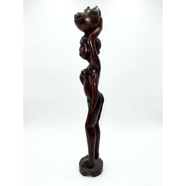 A striking and commanding 35-inch tall hand-carved solid hardwood sculpture depicting an African woman carrying a...