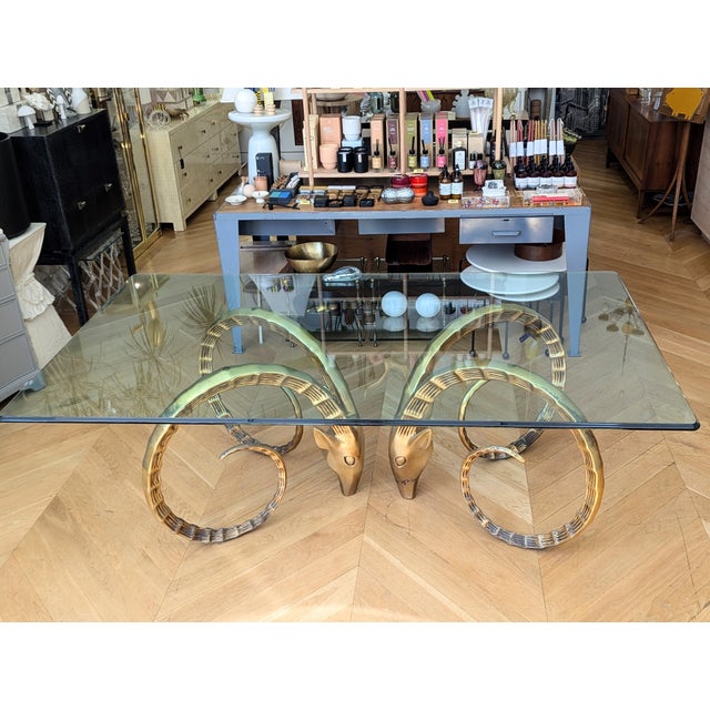 A striking vintage Ibex dining table, featuring sculptural brass Ibex bases and a sleek glass top. This Hollywood Regency-...