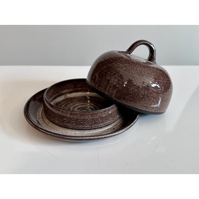 A round, handmade butter dish from the Serocka workshop, crafted in a brown, beige-streaked earthenware glaze on a dark...