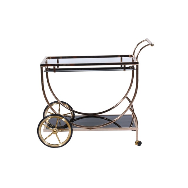 A brushed brass trolley or bar cart with glass shelves. Handle on once side, larger wheels on the other and swivel wheels...