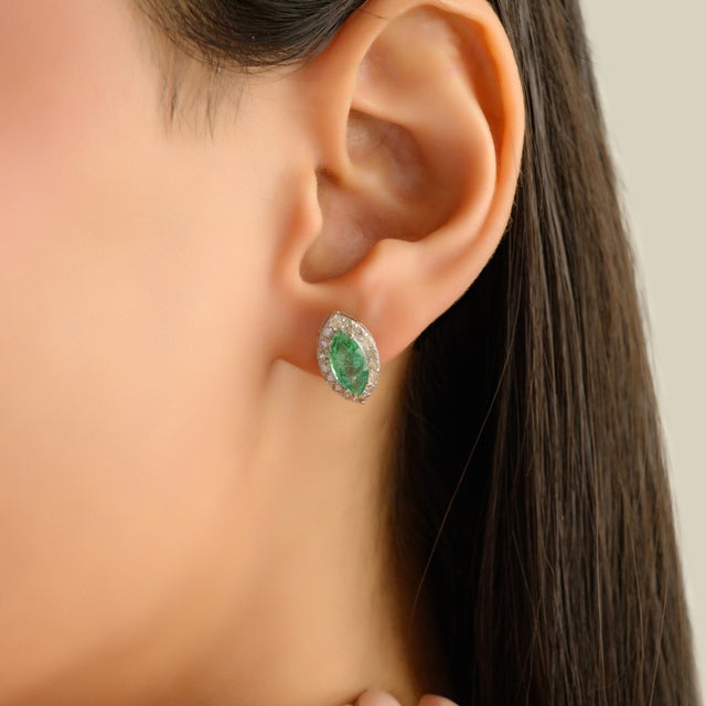 Striking silver stud earrings featuring marquise-cut emeralds totaling approximately 1.80 carats, framed by a delicate...