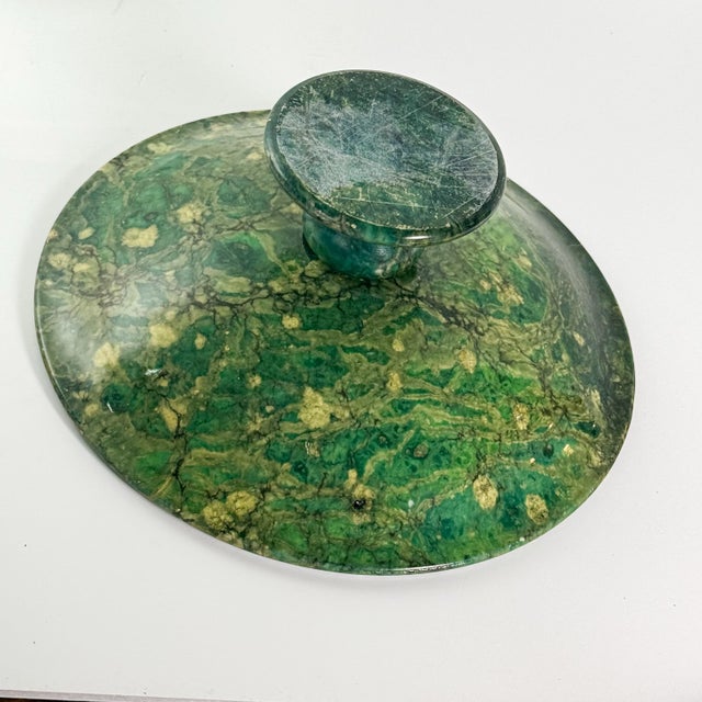 A shallow green stone bowl raised on a turned pedestal foot, with richly varied tones of deep green, teal, and cream...