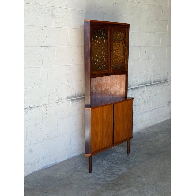 This display cabinet features a space-saving corner profile with patterned glass upper doors and solid lower doors. An...