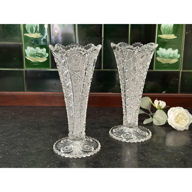 A matching pair of vintage clear glass trumpet vases featuring intricate pressed-glass patterns, scalloped rims, and...