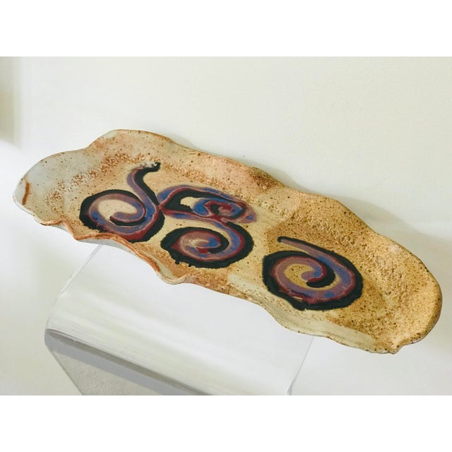 A glazed stoneware tray of asymmetric form. The subtle earth tones provide a background for the expressive color swirls of...