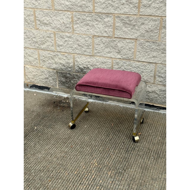 Vintage 1970s vanity stool with a clear lucite frame, brass accents, and pink velvet upholstery. Simple curved legs with...
