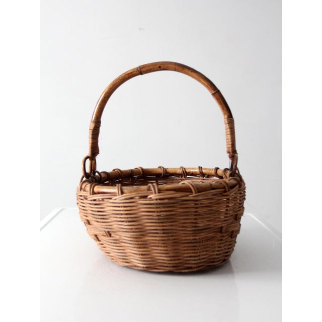 A vintage woven gathering basket with a bent bamboo handle, oval form, and tightly woven wicker body in rich honey brown...