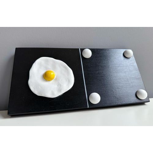 Mixed-media wall panel with foam clay fried egg motif on black lacquered wood domino panel combining playful humor...