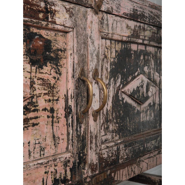 A striking wooden shelf cabinet defined by its dramatic, time-formed patina in soft blush pink layered beneath deep...