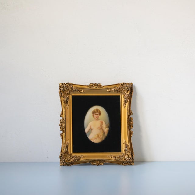 An antique hand-painted porcelain plaque depicting a classical female nude holding a strand of thread, presented in an...