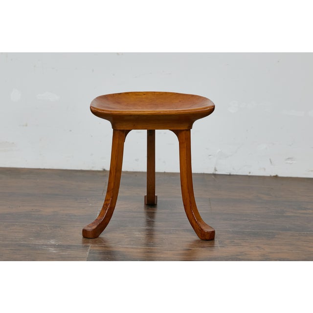 An English Liberty & Co. walnut Thebes stool from the Turn of the Century with tripod base. In-curving wooden seat resting...