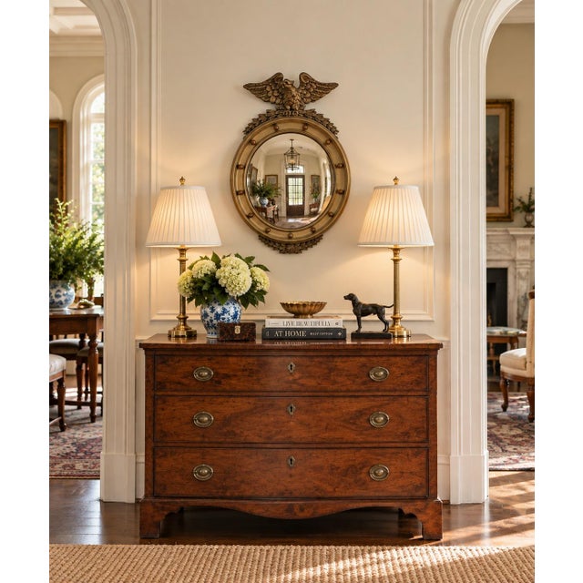 A Federal-style giltwood convex mirror crowned with a carved eagle, its wings outstretched above a laurel motif—an...