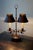 A sculptural double-arm table lamp in darkened forged iron, fitted with pierced conical tole shades and candle-style...