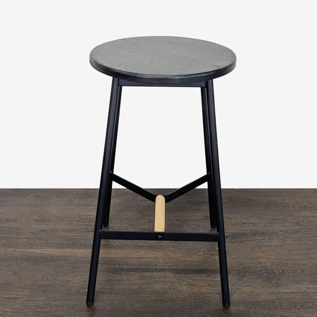 This round stool features a sleek black metal frame with a matching black wooden seat, complemented by a natural wood...
