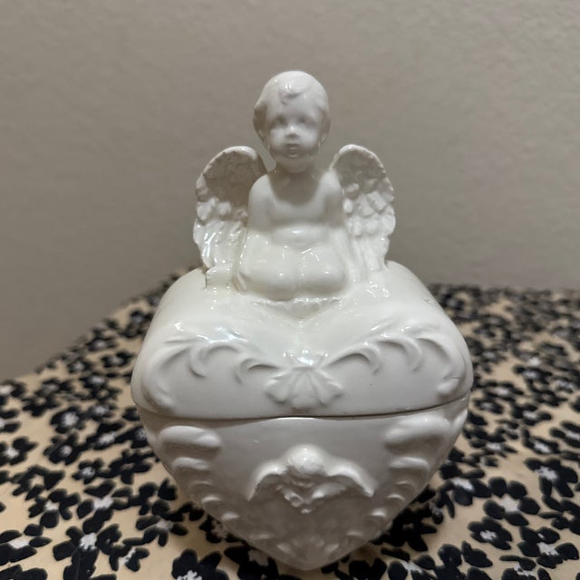 This charming vintage trinket box features a seated cherub (or angel) atop a heart-shaped lid. Crafted from elegant white...