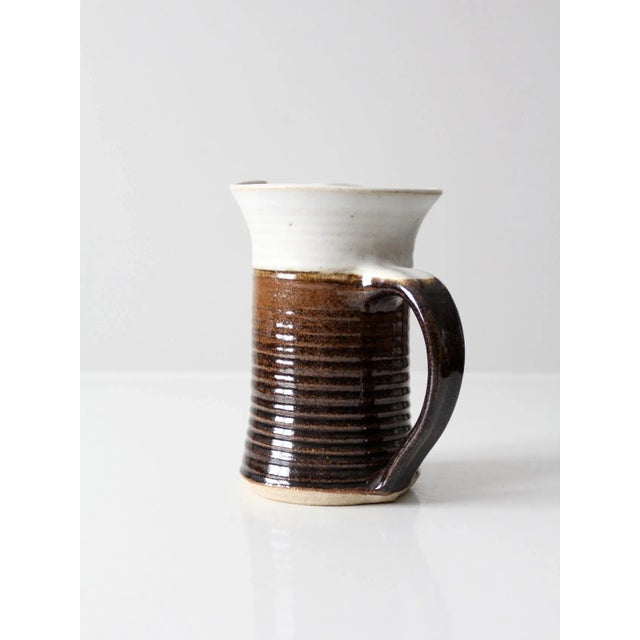 This vintage signed studio pottery pitcher is a beautifully handcrafted ceramic piece featuring a warm brown and white...