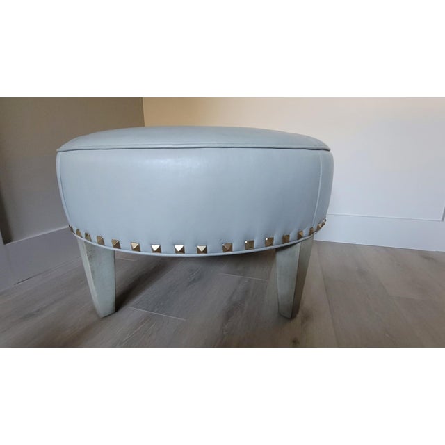 Two Dove grey round leather ottomans with modern square raised brass nailhead/rivet detail. Legs have rubbed subtle grey...