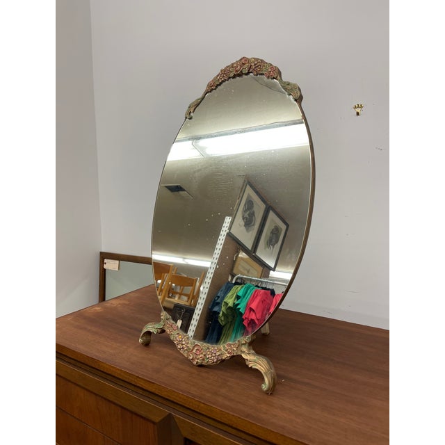 This vintage oval vanity mirror features a decorative frame adorned with hand-painted floral motifs in soft tones of...