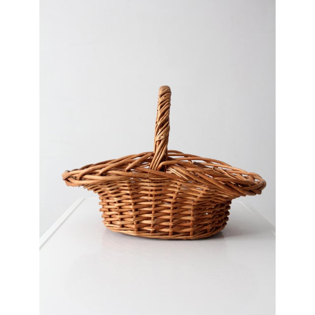This vintage handwoven wicker gathering basket features a generous oval shape and softly arched handle, with a woven...